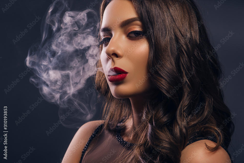 Naklejka premium Glamour caucasian brunette woman in evening black dress on a neutral background. She pose sensually, blowing smoke from her mouth. Retro fashion mood.
