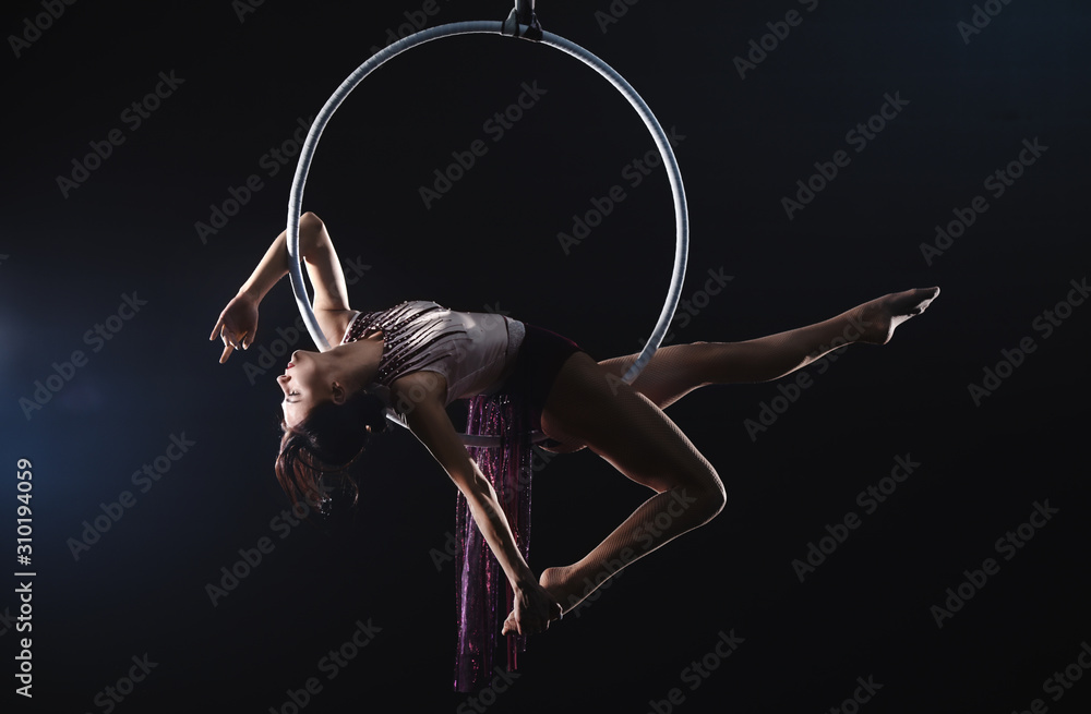 Fototapeta Young woman performing acrobatic element on aerial ring against dark background