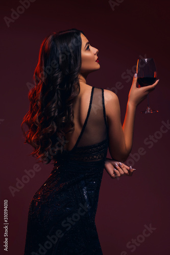Glamour caucasian brunette woman in evening black dress on a neutral background. She pose sensually with glass of wine. Retro fashion mood.