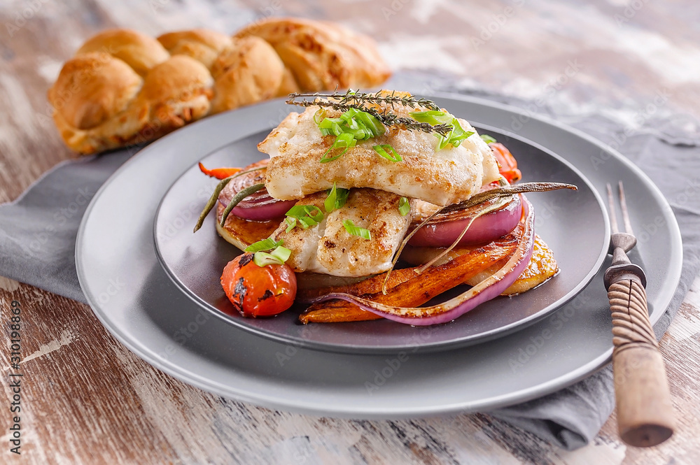 Closeup appetizing cod filet with red onion and herbs, baked potatoes and carrots on a side dish in a gray plate on a napkin. Tasty and healthy seafood