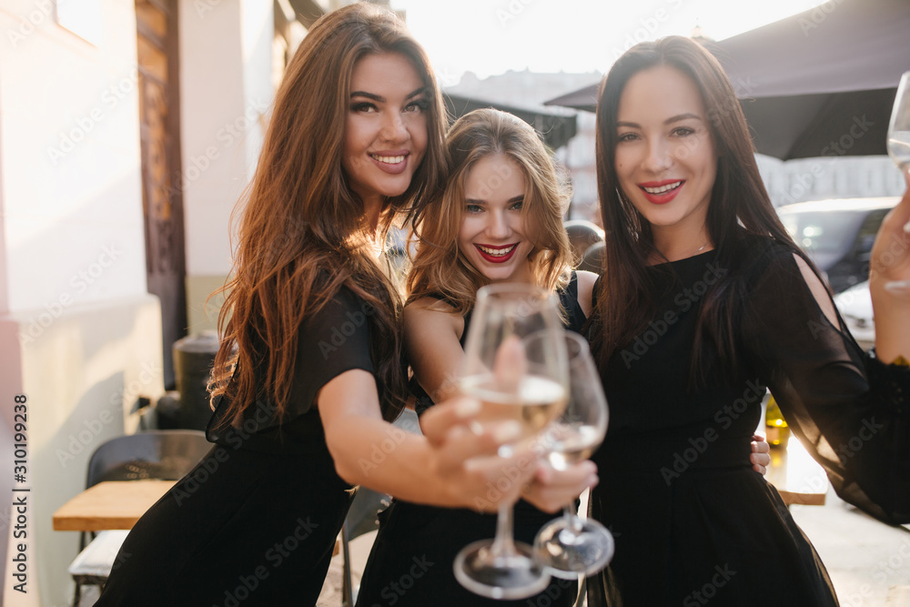 Pretty joyful girl with long beautiful hair raises glasses with her friends. Outdoor portrait of young ladies with trendy make-up wears black dresses at birthday party. Pretty joyful girl with long beautiful hair raises glasses with her friends. Outdoor portrait of young ladies with trendy make-up wears black dresses at birthday party.