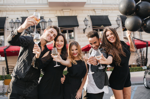 Blissful lady with long curly hair clink wineglasses with friends during photoshoot beside restaurant. Attractive female model in black dress celebrating birthday and enjoying outdoor party.