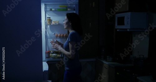 A woman opens a refrigerator at night and takes a bottle of water