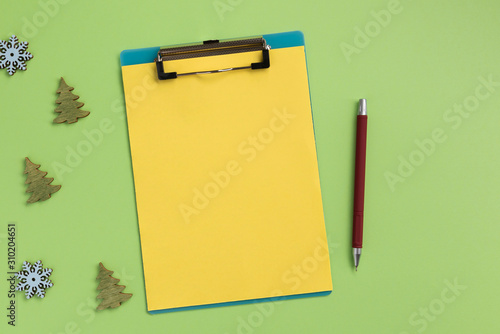 Clipboard with yellow sheet and christmas decor, on green background. Flat lay, top view, copy space.