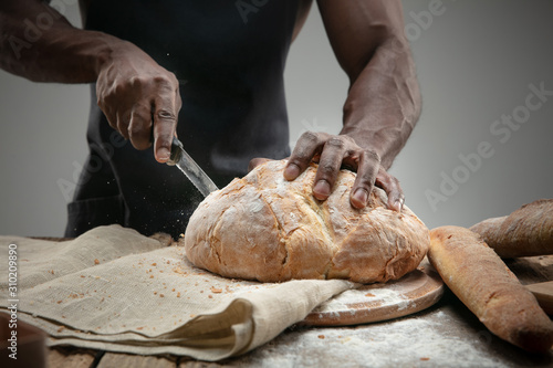 Close up of african-american man slices fresh cereal, white bread, bran with a kitchen knife on wooden table. Healthy eating, nutrition, craft product. Gluten-free food, vegan lifestyle, organic taste