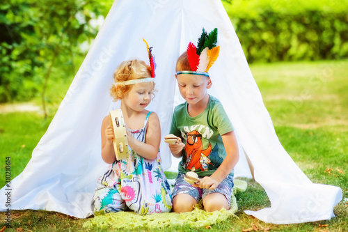 3 years old girl and 5 years old boy with feathers on head sit near white tent with tambourine and maracas in hands. Children pretend to be indian. Indians game in summertime. Children play music photo