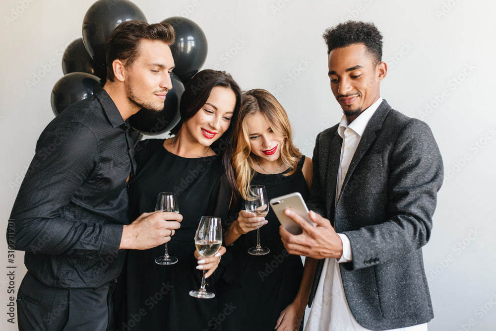 Confident african man in stylish gray jacket making selfie on event with colleagues. Pretty blonde girl looks with interest at phone screen during birthday party with best friends. Confident african man in stylish gray jacket making selfie on event with colleagues. Pretty blonde girl looks with interest at phone screen during birthday party with best friends.