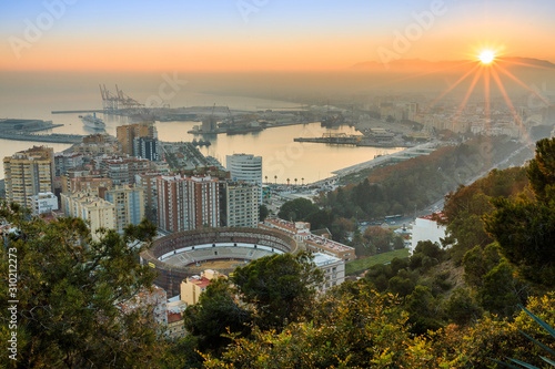 Sunset over Malaga. The city on the Spanish Costa del Sol in the evening mood with a panoramic view of the harbor, houses, trees, bullring with illuminated street lamp and blue sky over the mountains