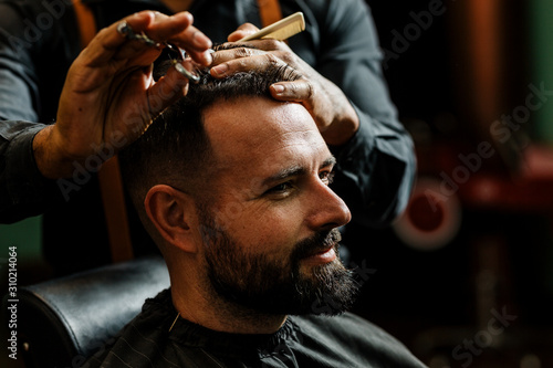 un barbero afroamericano brasileño esta cortando con tijeras y peine el pelo de la cabeza (cuero cabelludo, cabello) a un hombre caucásico con barba. de estilo hipster, vintage. photo