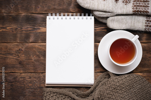 Blank notepad with copy space, warm clothes and cup of tea on brown wooden table flat lay background.