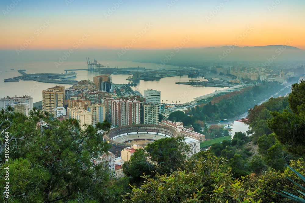 Obraz premium Evening sun over the Spanish city of Malaga on the Mediterranean coast. View of the city center on the Costa del Sol with illuminated buildings, trees, street lamps, harbor, ships