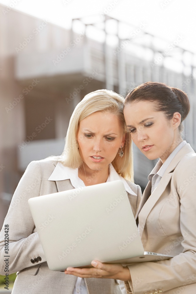 Young serious businesswomen working on laptop while standing against office building Young serious businesswomen working on laptop while standing against office building