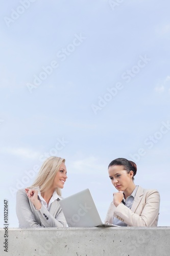 Low angle view of concentrated businesswoman using laptop while standing with coworker on terrace against sky