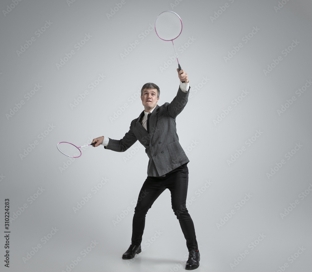 Time for emotions. Man in office clothes plays badminton with two racket on grey background. Businessman training in motion, action. Unusual look for sportsman, activity. Sport, healthy lifestyle. Time for emotions. Man in office clothes plays badminton with two racket on grey background. Businessman training in motion, action. Unusual look for sportsman, activity. Sport, healthy lifestyle.