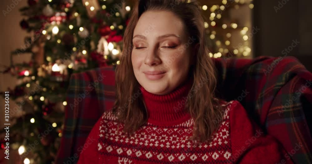 Close up of beautiful girl in a red sweater sits in armchair near Christmas tree with closed eyes waiting for a surprise. She opens his eyes and enjoys the gift. Dolly zoom shot Close up of beautiful girl in a red sweater sits in armchair near Christmas tree with closed eyes waiting for a surprise. She opens his eyes and enjoys the gift. Dolly zoom shot