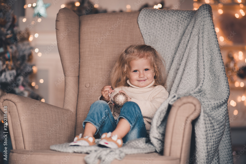 Pretty baby girl 4-5 year old wearing knitted sweater and denim pants sitting in chair over Christmas lights close up. Looking at camera. Childhood. Pretty baby girl 4-5 year old wearing knitted sweater and denim pants sitting in chair over Christmas lights close up. Looking at camera. Childhood.
