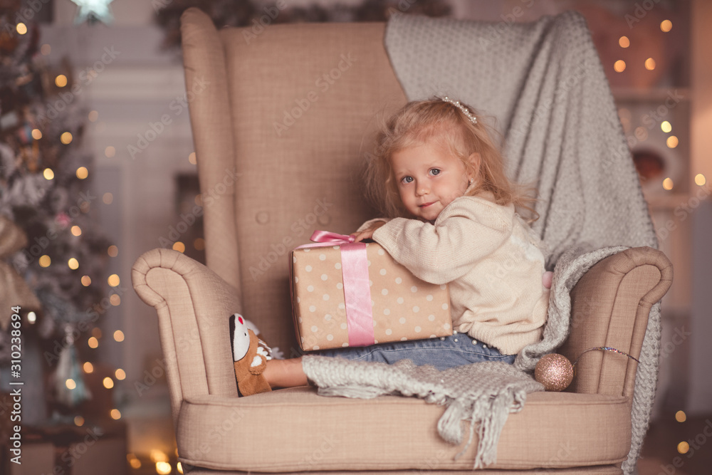 Funny baby girl 3-4 year old open Christmas present sitting in chair over xmas lights closeup in room. Looking at camera. New Year. Winter holiday season. Funny baby girl 3-4 year old open Christmas present sitting in chair over xmas lights closeup in room. Looking at camera. New Year. Winter holiday season.