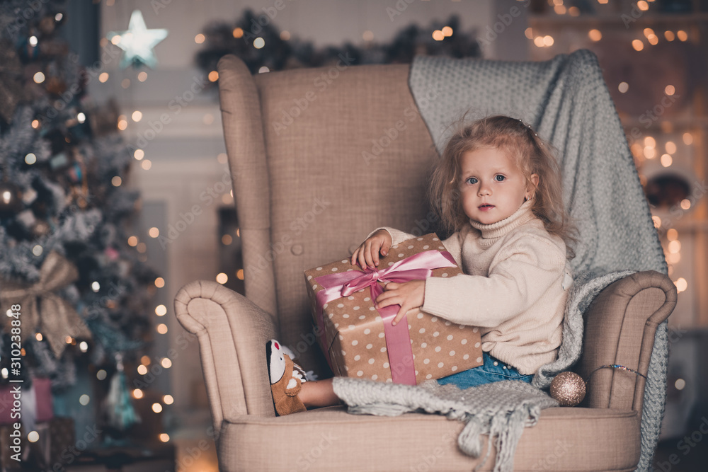 Cute baby girl 3-4 year old open Christmas presents sitting in armchair over glowing lights closeup. Looking at camera. New Year. Childhood.