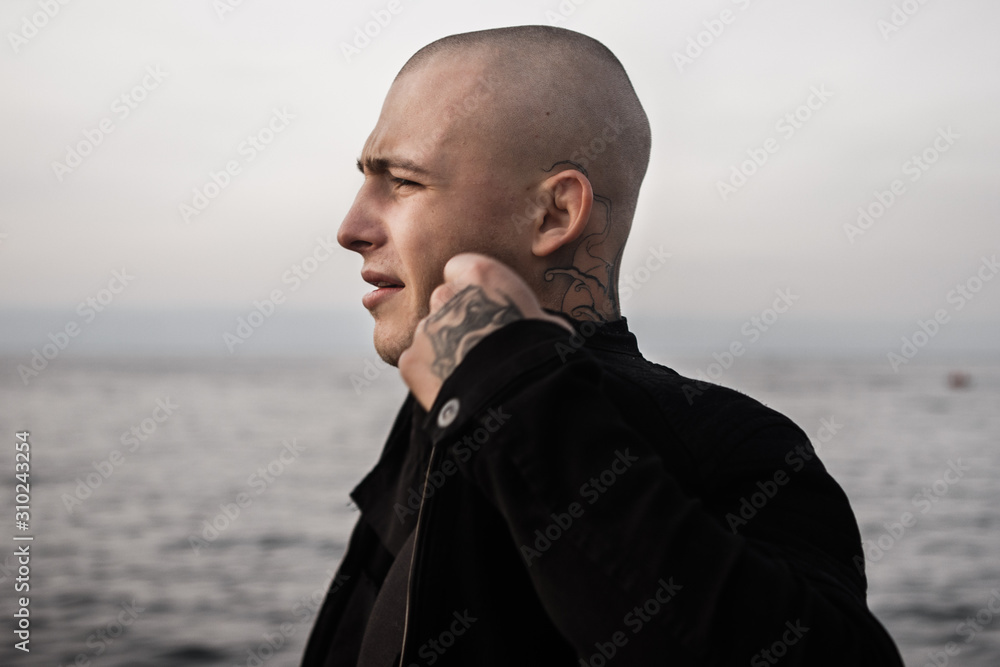 Brutal tattooed guy in a black denim jacket spreads his collar and looks thoughtfully into the distance at sea. A guy with tattoos on the ocean. Style and fashion, concept of bald bad hooligan guy. Brutal tattooed guy in a black denim jacket spreads his collar and looks thoughtfully into the distance at sea. A guy with tattoos on the ocean. Style and fashion, concept of bald bad hooligan guy.