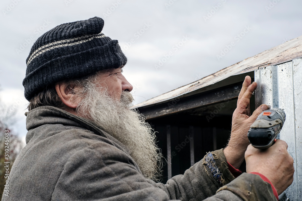 An elderly gray bearded man in a headdress and a worn jacket is engaged in construction work in his yard An elderly gray bearded man in a headdress and a worn jacket is engaged in construction work in his yard
