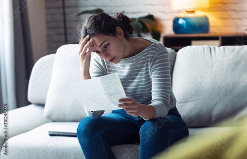 Cuadro en lienzo Worried young woman looking at bills and thinking about her problems while sitting on the sofa at home