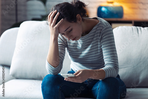 Worried young woman using her mobile phone while sitting on sofa in the living room at home. photo