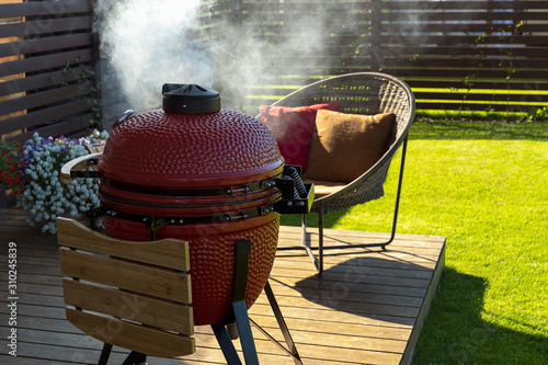 Barbecue grill and rattan armchair in cozy home terrace. Smoke coming out of red ceramic Barbecue. 