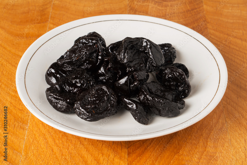 Dried black plum fruits (prunes) in a white plate on a wooden work Dried black plum fruits (prunes) in a white plate on a wooden work
