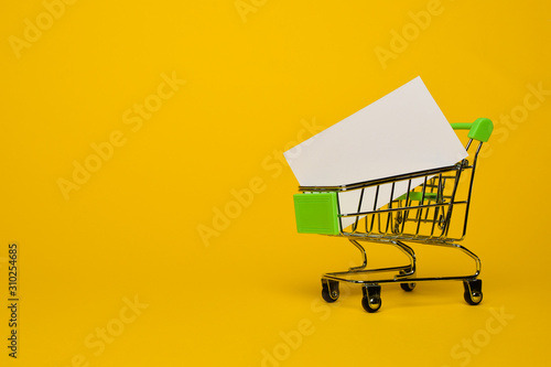 a small metal trolley from a supermarket carries a white banner on a yellow background. Horizontal photo. Place for text. Trade concept