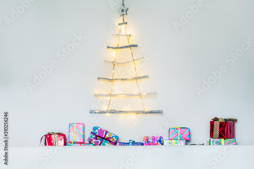 Modern creative christmas eco tree made of wooden sticks hanging on white wall with festive lights and colourful gift boxes. Simple, minimal conscientious interior design and decor. Copy space. © okrasiuk