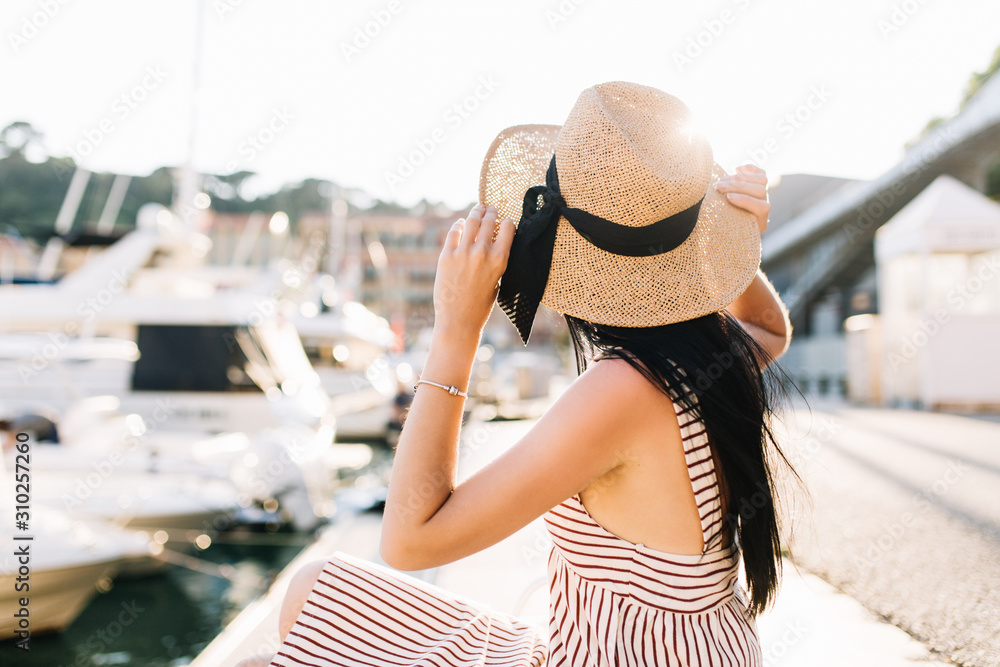 Stylish graceful woman in hat with ribbon looking at boats and yachts resting in seaport in summer day. Outdoor portrait of tanned brunette girl enjoying vacation in Europe and chilling outside.