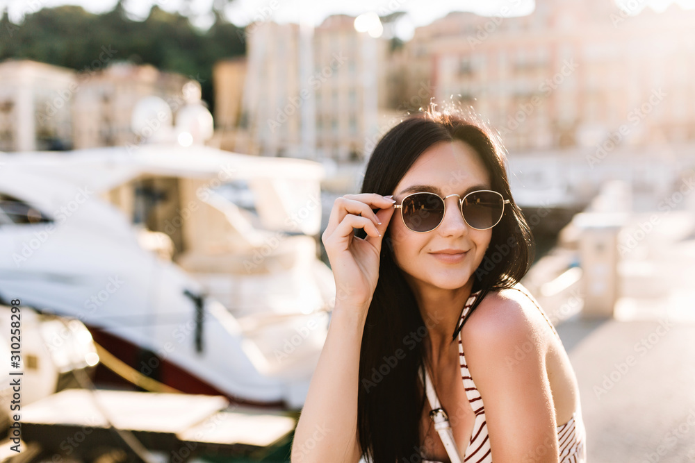 Attractive glad girl with bronze skin holding black sunglasses and resting outdoor with boats on background. Portrait of chilling brunette young woman enjoying weekend on resort and sunbathing.