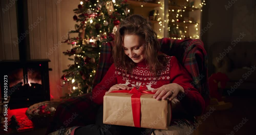 Happy emotional girl with a gift in her hands near the Christmas tree in decorated beautiful a cozy country house. Holiday christmas spirit. Opening a gift Happy emotional girl with a gift in her hands near the Christmas tree in decorated beautiful a cozy country house. Holiday christmas spirit. Opening a gift