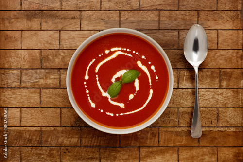 A bowl of delicious tomato soup, topped with a drizzle of cream and a sprig of basil, in a rustic bowl, shot on a wooden background, with a spoon.