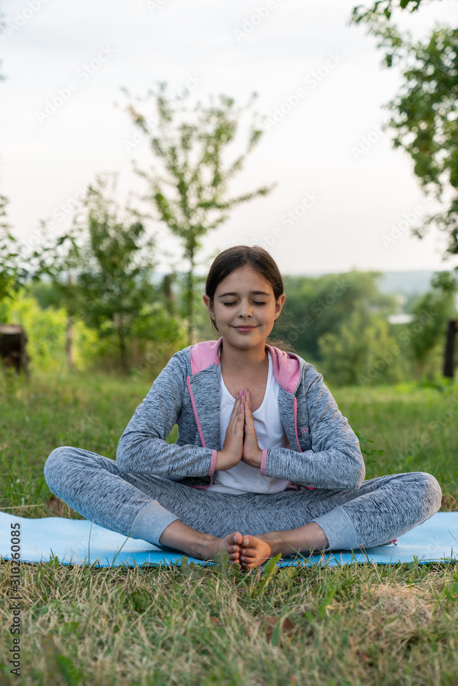 Outdoor front view full body portrait of a relaxed brunette girl in grey sportswear practicing yoga sitting on the yoga mat among the garden Outdoor front view full body portrait of a relaxed brunette girl in grey sportswear practicing yoga sitting on the yoga mat among the garden