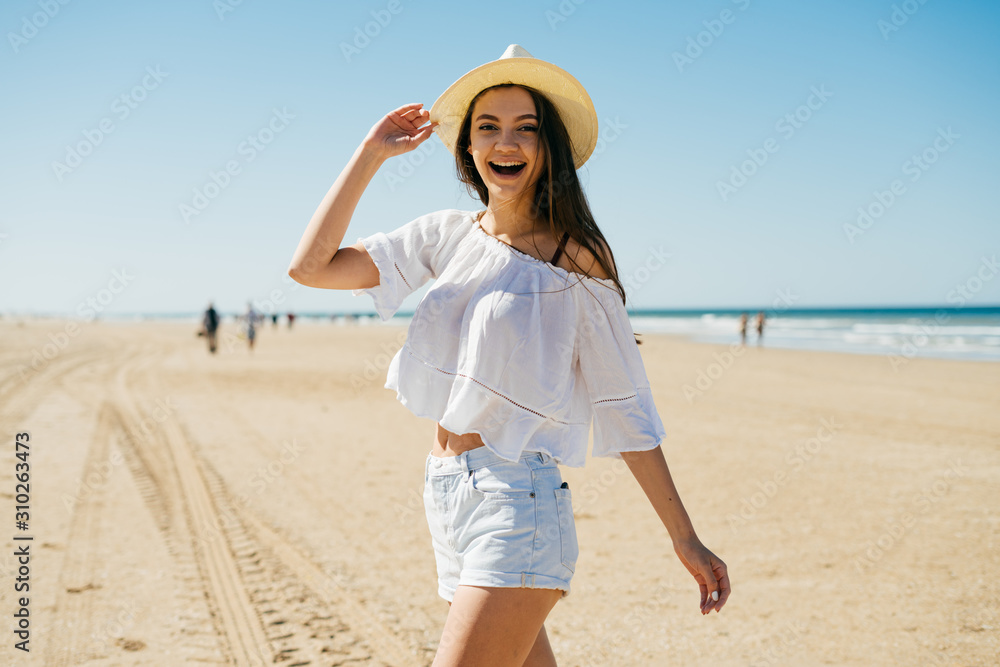 girl dynamically walking along the beach past the camera, laughs, holding her hat with her hand girl dynamically walking along the beach past the camera, laughs, holding her hat with her hand