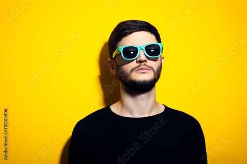 Studio portrait of young serious man wearing cyan sunglasses and black sweater on yellow background.