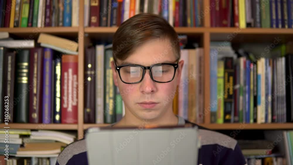 A young man is reading a book in a tablet. A man with glasses carefully looks at the tablet. In the background are books on bookshelves. Book library. A young man is reading a book in a tablet. A man with glasses carefully looks at the tablet. In the background are books on bookshelves. Book library.