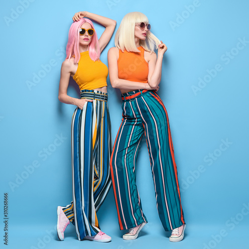 Two fashionable girl posing in Studio. Beautiful Adorable young woman in Stylish summer colorful outfit, Trendy dyed hair, make up. Glamour model sister friend, fashion concept on blue, full-length
