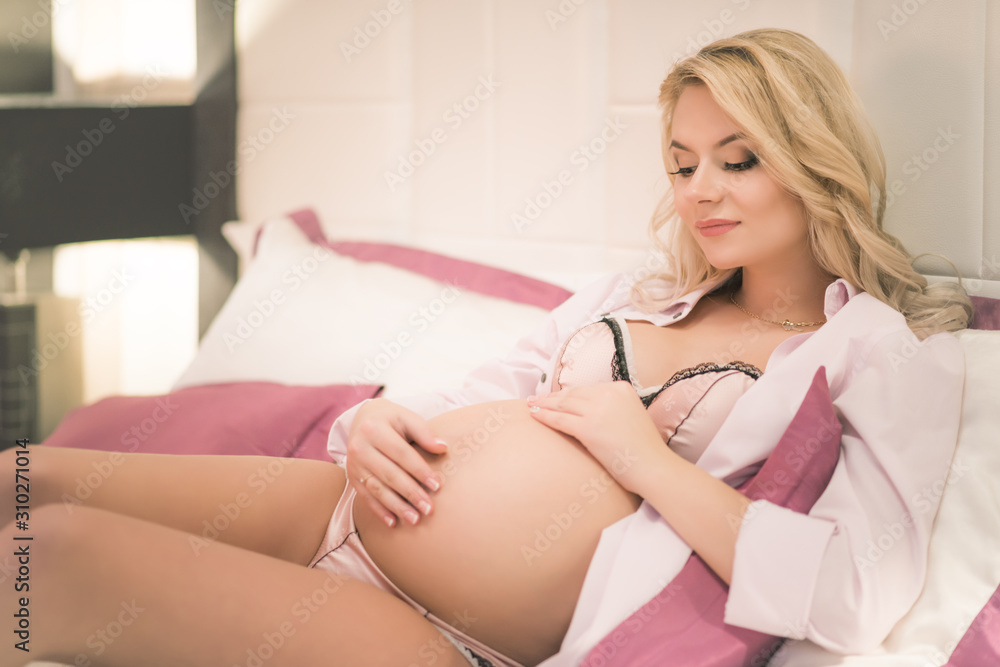 Young beautiful woman with a pregnant belly stroke her stomach, rejoices to her newborn child sitting in a shirt and underwear in a beautiful interior Young beautiful woman with a pregnant belly stroke her stomach, rejoices to her newborn child sitting in a shirt and underwear in a beautiful interior