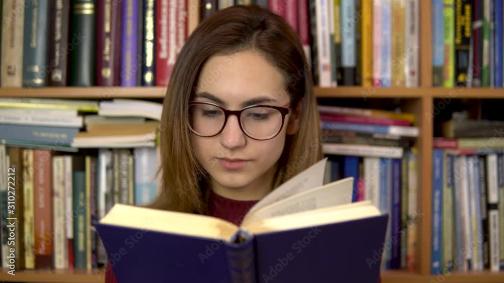 A young woman is reading a book in a library. A woman with glasses carefully looks at the book closeup. In the background are books on bookshelves. Book library. A young woman is reading a book in a library. A woman with glasses carefully looks at the book closeup. In the background are books on bookshelves. Book library.
