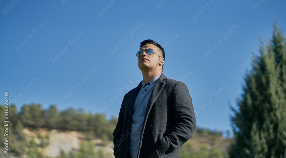 portrait of a handsome Hispanic boy standing on the stairs with good pants and urban clothes: jeans, blue shirt and dark jacket and sunglasses on his face. blue background portrait of a handsome Hispanic boy standing on the stairs with good pants and urban clothes: jeans, blue shirt and dark jacket and sunglasses on his face. blue background