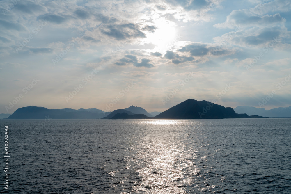 Sun setting over backlit Islands in the Fjords of Norway with calm waters and clouds in the sky from a boats perspective Sun setting over backlit Islands in the Fjords of Norway with calm waters and clouds in the sky from a boats perspective