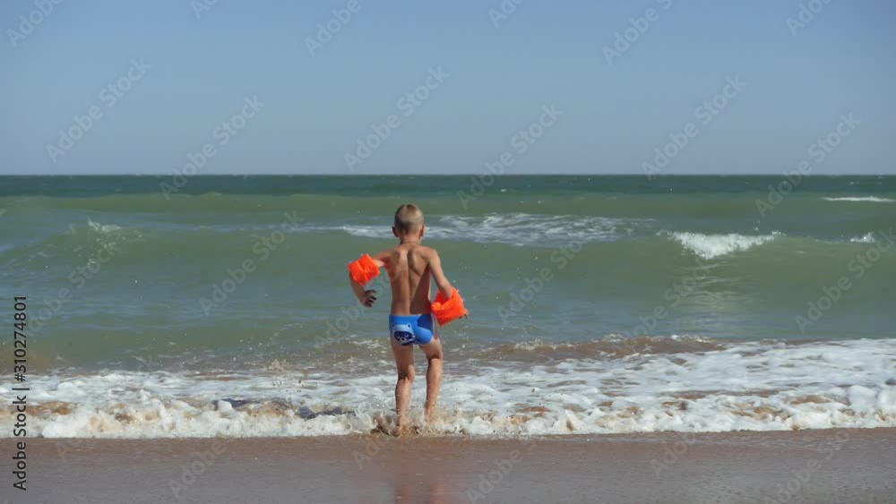 Boy runs into the sea. Waves wash over the beach. Child in armbands on vacation at sea. The child dives into the raging waves. Boy runs into the sea. Waves wash over the beach. Child in armbands on vacation at sea. The child dives into the raging waves.
