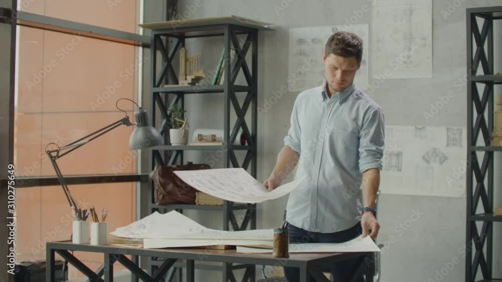 A male architect looks at drawings and plans of buildings in a loft office against a large window. The designer plans the facade of buildings. Creating furniture design. A male architect looks at drawings and plans of buildings in a loft office against a large window. The designer plans the facade of buildings. Creating furniture design.