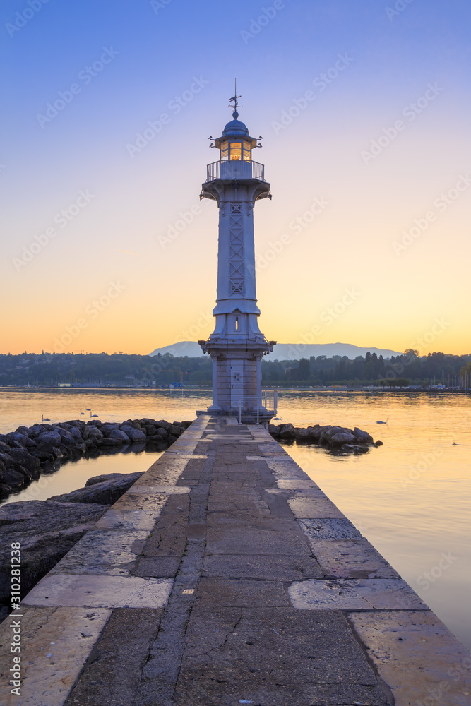 Stone walkway and rocks at the end of the Paquis peninsula on Geneva lake early in the morning, before sunrise. Lighthouse is a silhouette, and orange hue on the horizon is reflected in calm water.