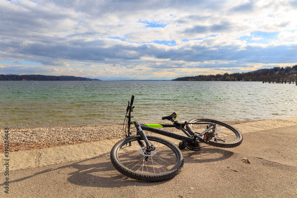 A bicycle is laying on the ground by Lake Starnberg in Bavaria, Germany, on a sunny day under dramatic sky with low clouds.