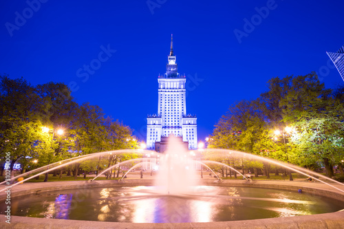 Public park and Palace of Culture and Science in Warsaw, Poland at night. Historical building in art deco style at dusk, under clear blue sky, with a large fountain in the foreground.