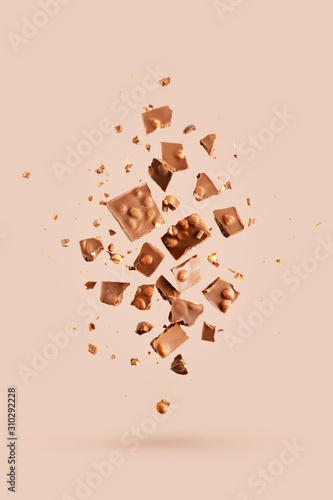 Flying in the air broken bar of milk chocolate with nuts and flakes on pastel pink background.  Chocolate pieces levitation concept.
