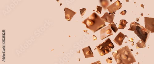 Flying in the air broken bar of milk chocolate with nuts and flakes on pastel pink background.  Chocolate pieces levitation concept. Wide banner.